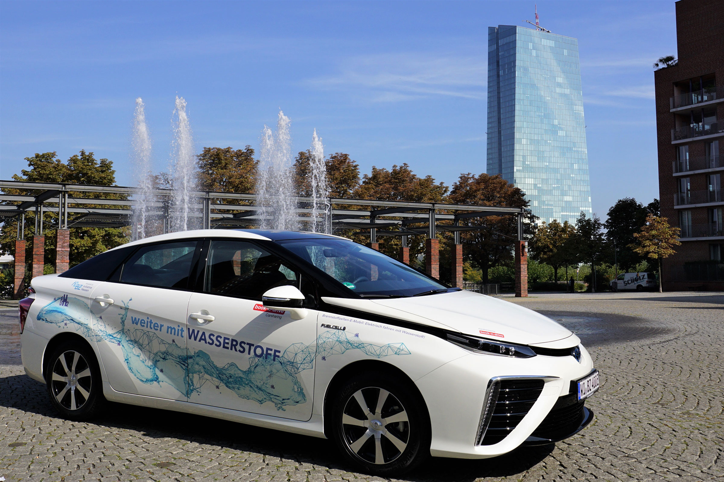 Weißer wasserstoffbetriebener Toyota Mirai der Wasserstoff- und Brennstoffzellen-Initiative Hessen e.V. steht in Frankfurt mit Springbrunnen und Gebäuden im Hintergrund.